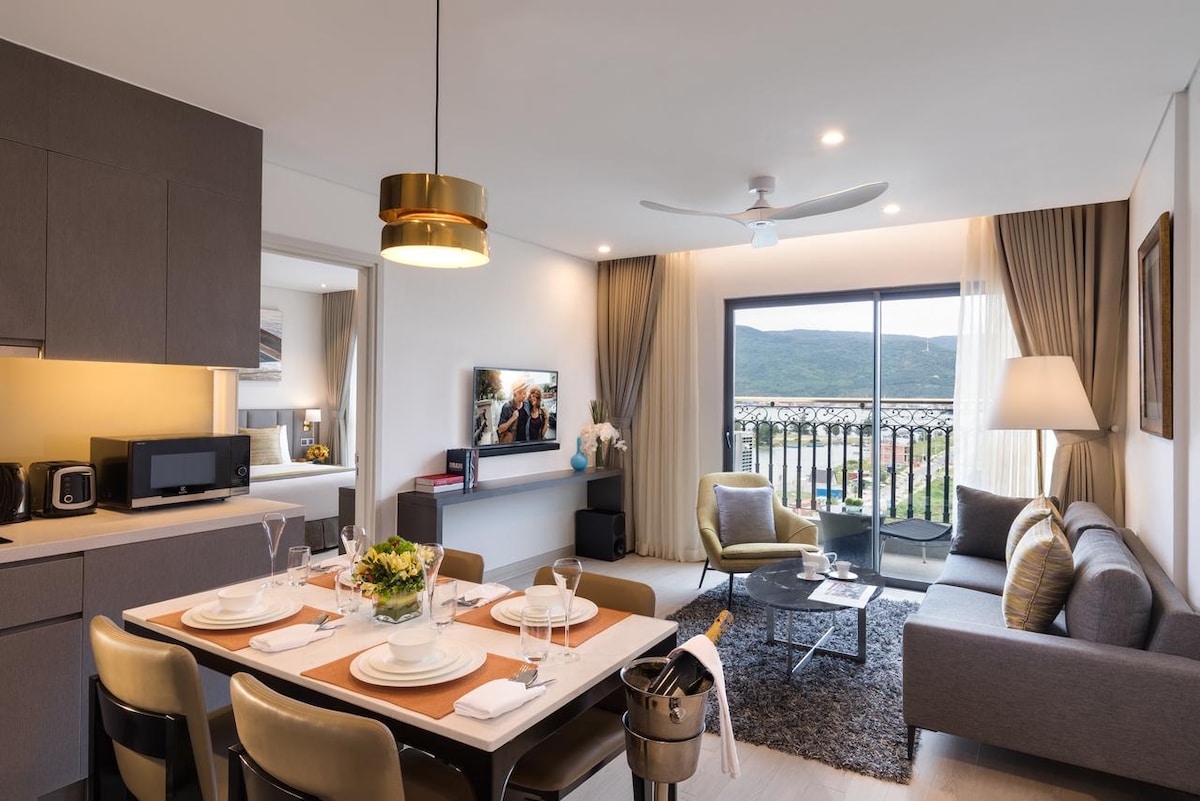 A modern living area is presented with a dining table set for four. A sleek kitchen is visible, featuring appliances like a microwave. A cozy seating area includes a sofa and a coffee table. Large windows offer views of the landscape beyond, complemented by soft curtains.