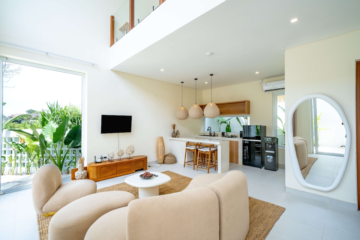 A modern living area features soft, rounded furniture arranged around a central table. The open kitchen is equipped with wood accents and pendant lighting. Large windows invite natural light and views of lush greenery, enhancing the spacious and inviting atmosphere.