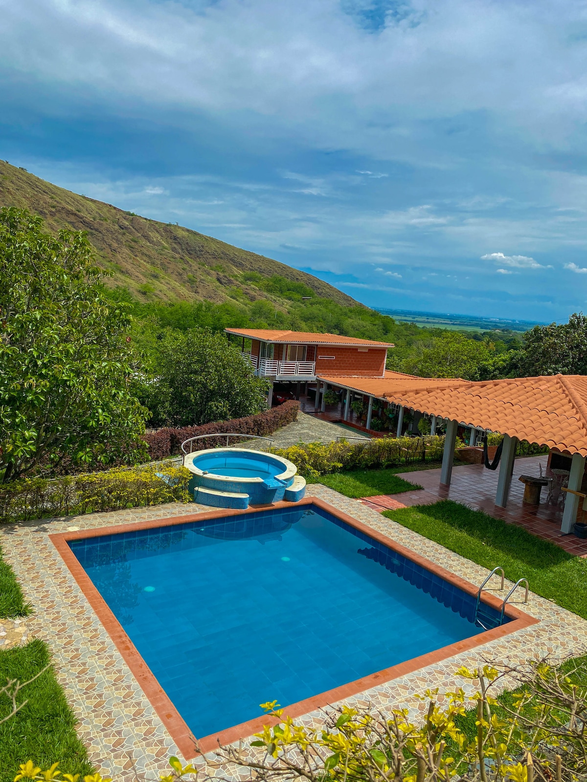 A spacious outdoor area features a large swimming pool surrounded by green landscaping. A circular jacuzzi is positioned adjacent to the pool. Nearby, a covered terrace provides shaded seating with a tan tiled roof, overlooking lush mountains and distant valleys.
