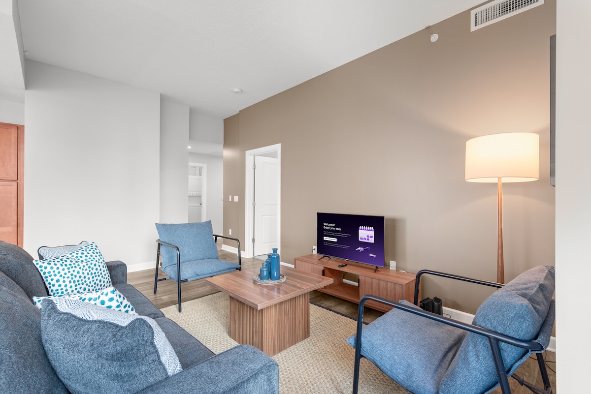 The living room features a comfortable sofa and two accent chairs, arranged around a wooden coffee table. A large flat-screen smart TV is mounted on the wall, with light streaming in from the nearby windows complemented by a floor lamp.