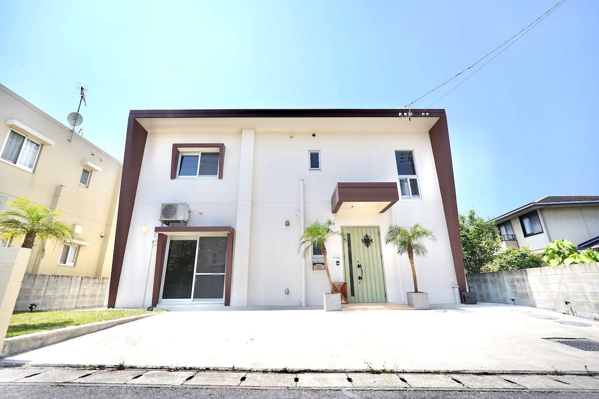 A modern two-story house is presented, featuring a minimalist facade with a symmetrical design. The entrance is adorned with a green door, framed by two small palm trees. The driveway is spacious and leads to a well-maintained exterior, under a clear blue sky.