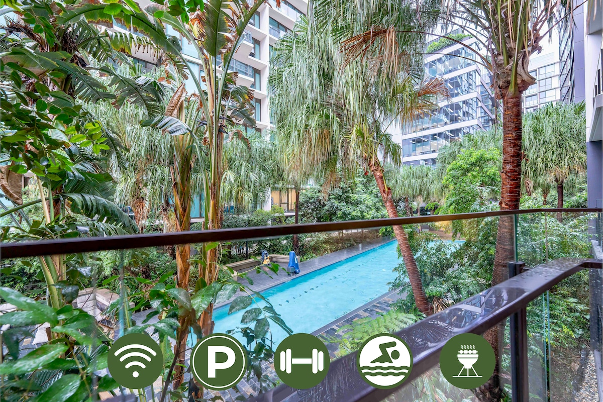 A balcony view reveals lush greenery surrounding a tranquil outdoor pool. Palm trees and other tropical plants provide a natural backdrop, creating a serene atmosphere. The pool's clear water is visible, inviting relaxation. Amenities include Wi-Fi, parking, a gym, and BBQ areas.