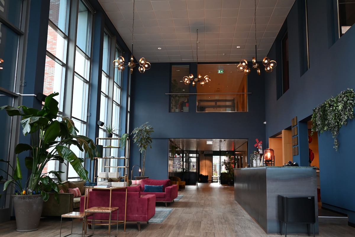 A spacious lobby area features large windows that allow natural light to fill the space. Stylish seating arrangements with colorful sofas are complemented by greenery from indoor plants. A modern reception desk stands at one side, creating an inviting atmosphere for guests.