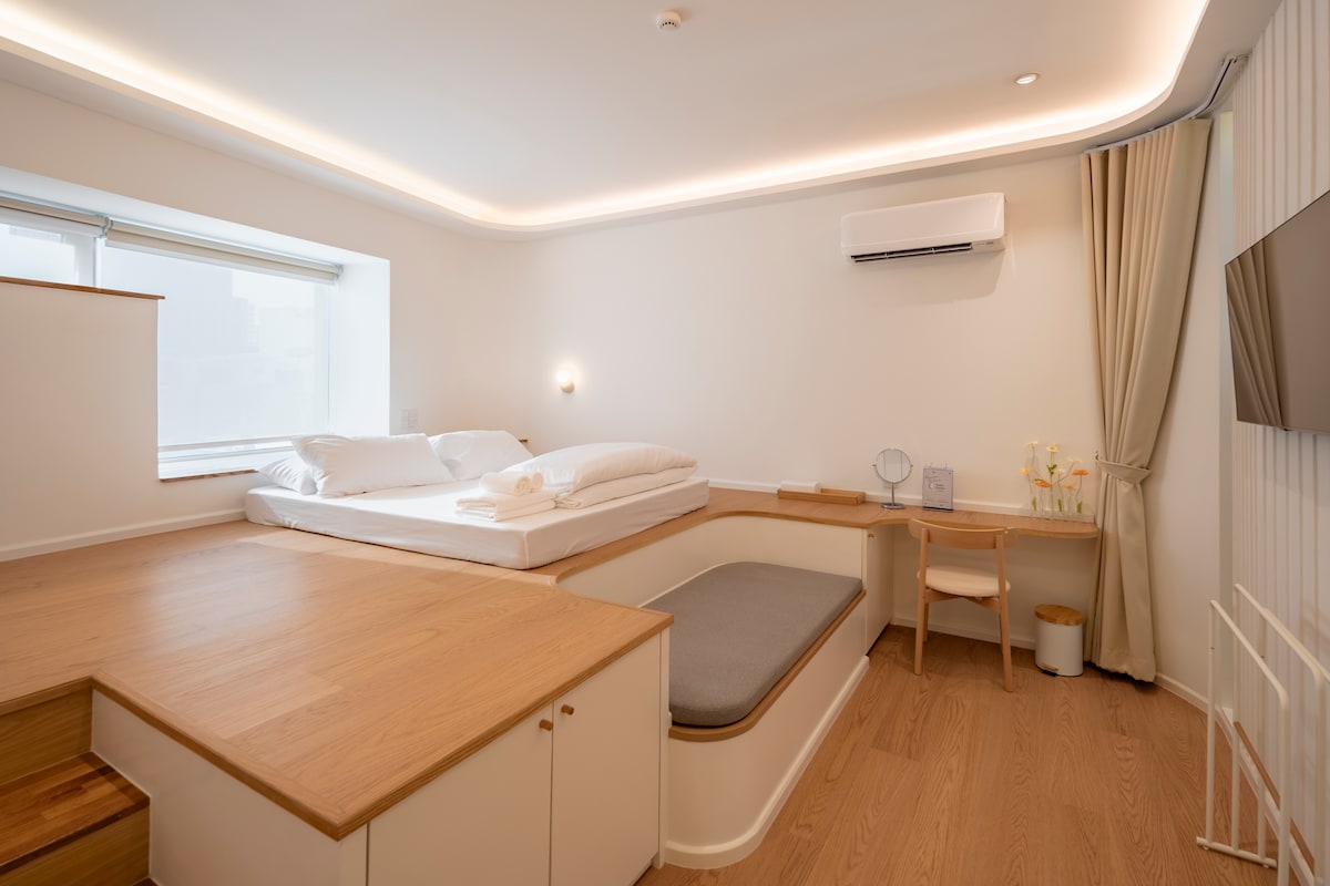 A bright room features stepped beds with crisp linens, a comfortable seating area, and minimalistic decor. A workspace is positioned beside a large window, providing natural light. Air conditioning is present, and a television is mounted on the wall for convenience.