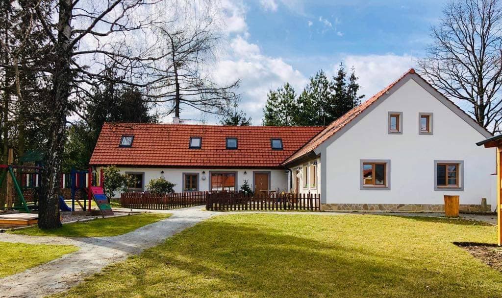 A spacious outdoor area features a well-maintained lawn, complemented by a bright white house with a red-tiled roof. The property includes a playground and a wooden fence outlining the garden, surrounded by trees and greenery.