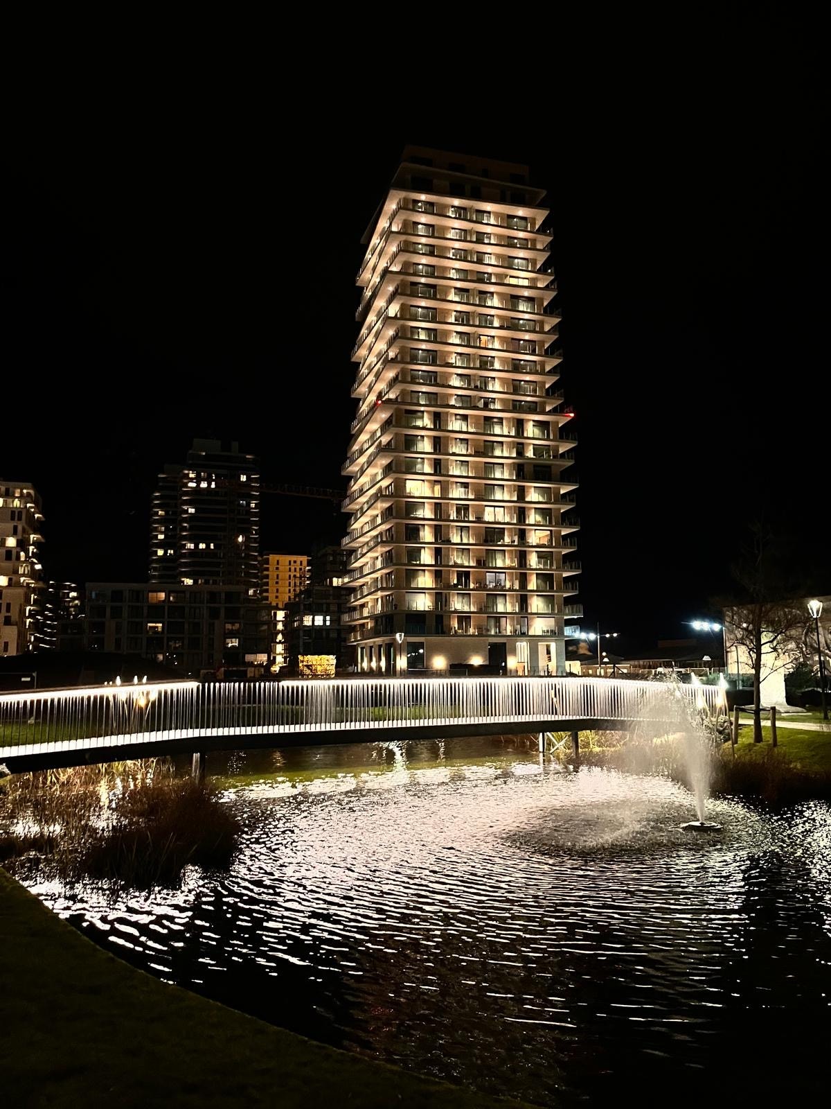 A modern building is illuminated against the night sky, showcasing a sleek design with numerous windows reflecting warm light. A small fountain is visible in the foreground, with water gently rippling in a pond surrounded by landscaped greenery. A pedestrian bridge arches over the water.