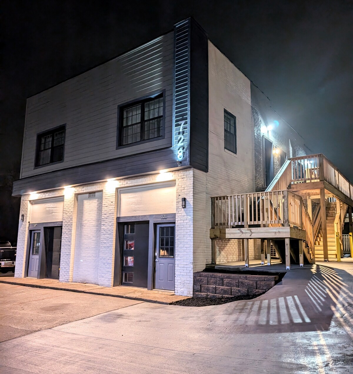 A modern, two-story building is illuminated at night, showcasing a sleek facade. The entrance features large, well-lit doors, while a wooden staircase leads to the upper level. Soft lighting highlights the clean lines of the structure against a dark sky.