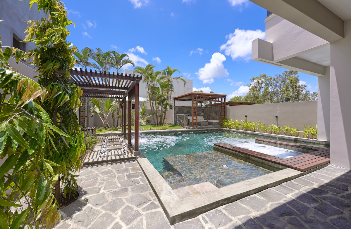 A luxurious pool area features a spacious jacuzzi and a cascade, surrounded by tropical greenery. Sunbeds are positioned under shaded pergolas, and natural stone tiles adorn the pool deck, creating a serene setting for relaxation.