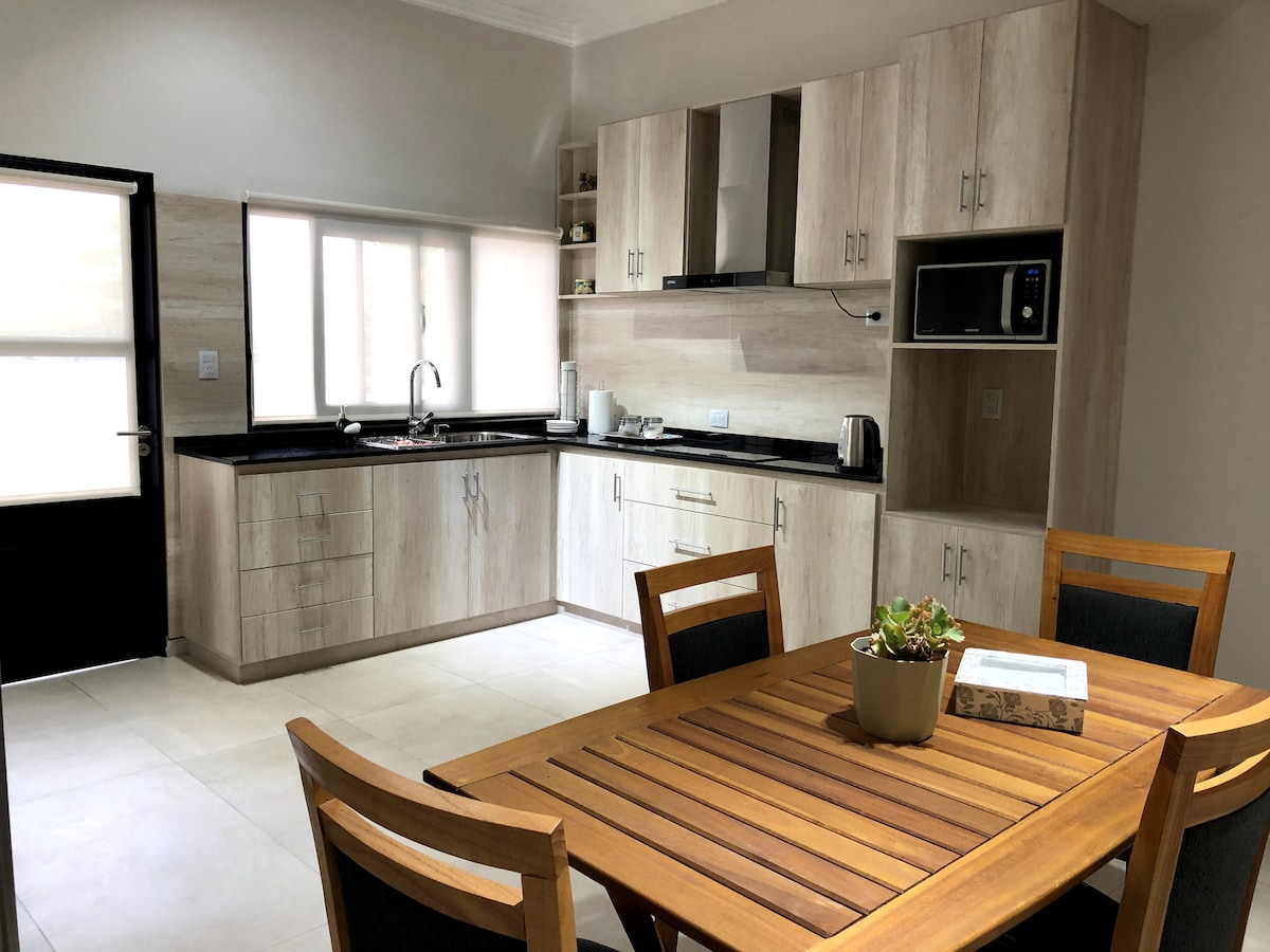 The kitchen features modern cabinetry with light wood finishes and ample counter space. A dining table with wooden chairs is positioned nearby. Natural light enters through the large window, highlighting the stainless steel appliances and minimalist decor.