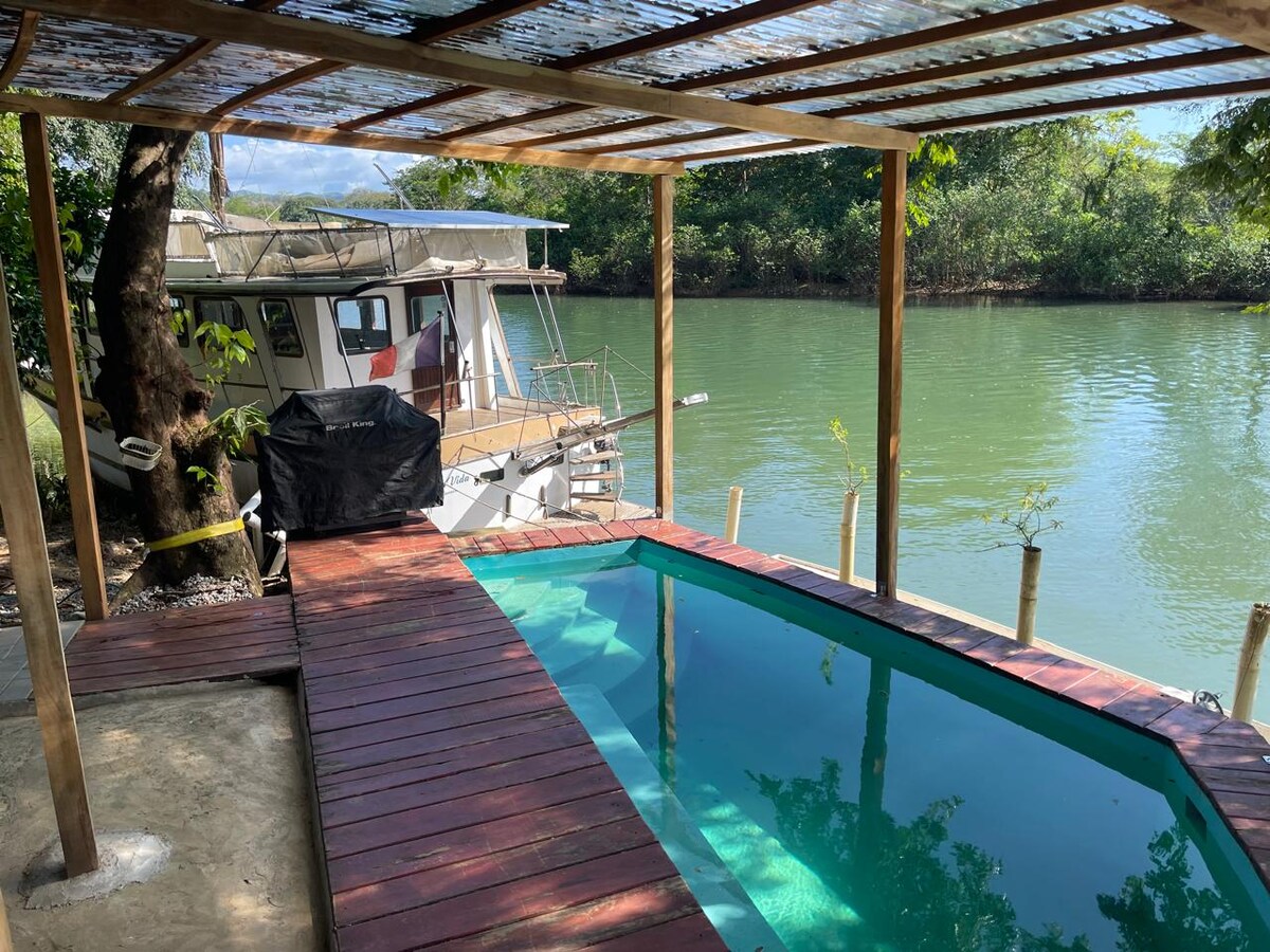 A private pool is framed by a wooden deck, offering a refreshing area beside calm green waters. A boat is visible nearby on the water, surrounded by lush vegetation and a clear blue sky.
