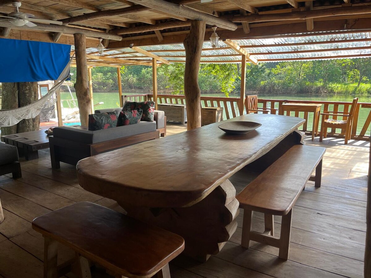 A large wooden dining table accommodates seating for several guests, complemented by additional wooden benches. Natural light filters through a translucent roof, illuminating the rustic decor. A hammock can be seen in the background, along with plush seating and green foliage visible outside.