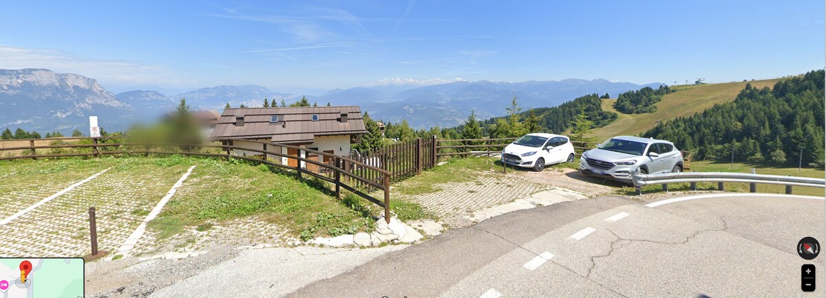 A scenic view of a parking area is presented, featuring two parked cars. The terrain slopes gently away from the building, with a wooden fence along the edge. Vibrant green grass and distant mountains create a natural backdrop under a clear blue sky.