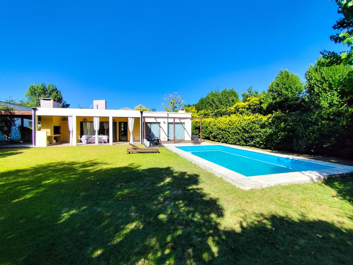 A spacious green lawn surrounds a private swimming pool, measuring 8 x 4 meters. The house features large windows that invite natural light, with a shaded area for relaxation. Lush trees and hedges create a tranquil atmosphere in the outdoor space.