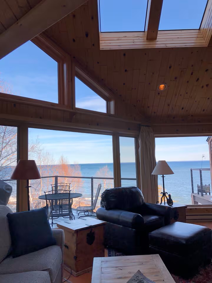 Large windows capture expansive views of Lake Superior and the surrounding landscape. Natural light fills the open living area, which features a comfortable seating arrangement with dark leather chairs and a cozy sofa. A small table set for two sits near the glass doors leading to the outdoors.