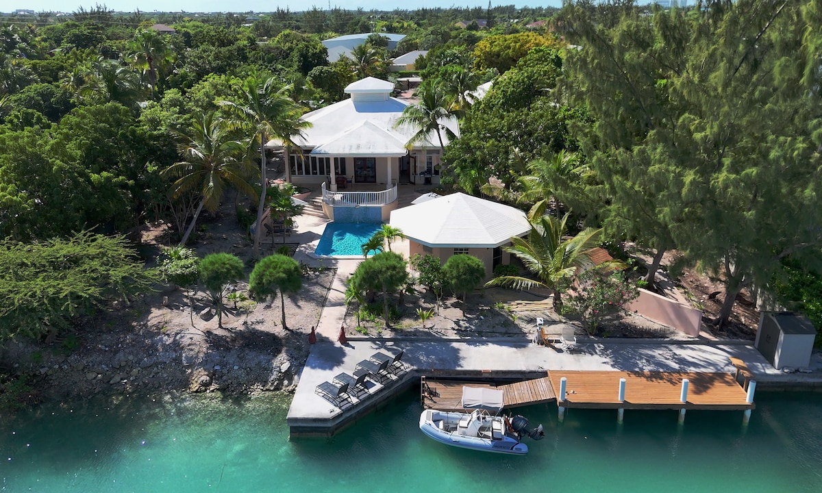 An aerial view of the canal-front villa showcases the distinct circular roofing of the main house along with lush landscaping. A private dock extends into the water, complementing the infinity pool that reflects the greenery surrounding it.