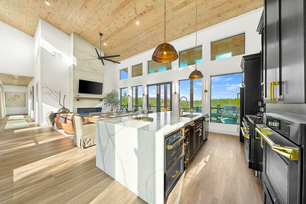 The kitchen showcases a spacious island with a sleek marble countertop, surrounded by modern black cabinetry. Large windows flood the area with natural light, offering views of the outdoors. Decorative pendant lights hang above, enhancing the elegant design of this culinary space.