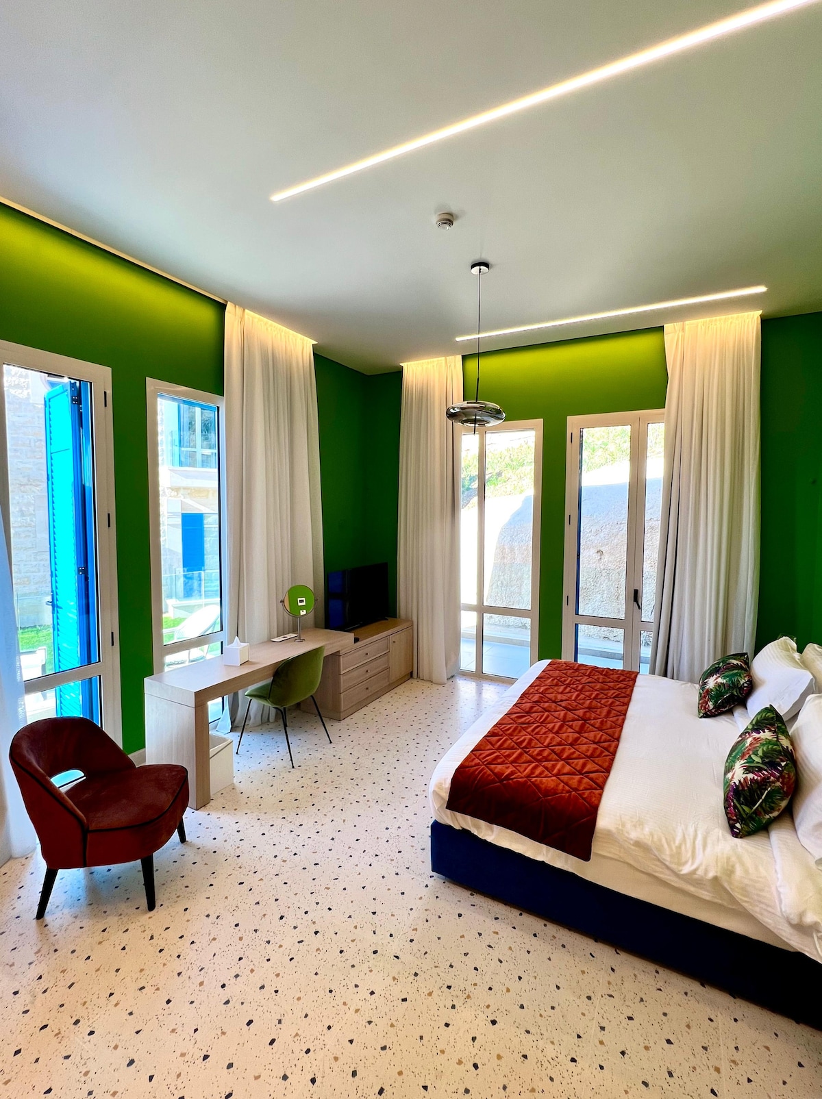 A spacious bedroom features vibrant green walls and large glass doors that open to natural light. A comfortable bed is adorned with decorative pillows. A desk and chair are positioned near a wall-mounted television, while soft drapes frame the windows, enhancing the airy ambiance.
