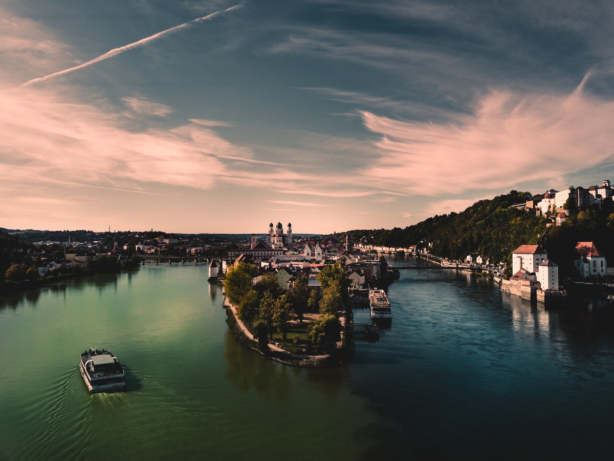 A scenic view of the river meandering through the landscape, bordered by lush greenery and historic buildings. Boats can be seen navigating the waters, while the sky is adorned with soft clouds, creating a serene atmosphere.