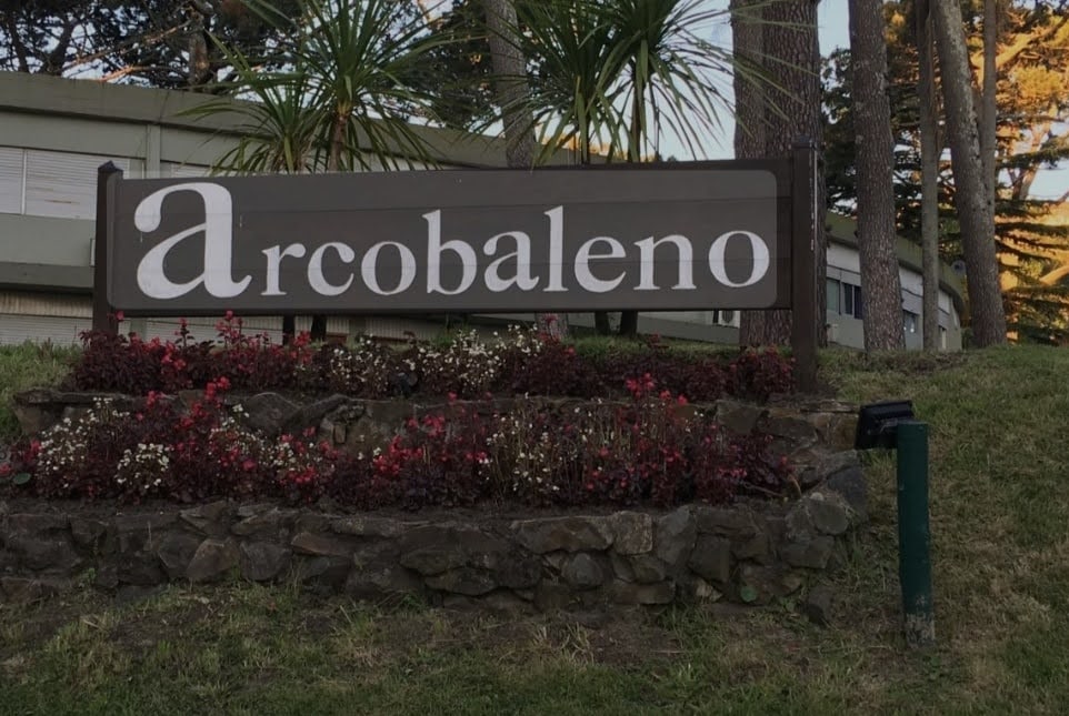 A sign reading 'Arcobaleno' is positioned at the entrance, surrounded by flowering plants and greenery. The sign is made of wood and features a clean, clear font, contributing to a welcoming aesthetic in the natural environment.