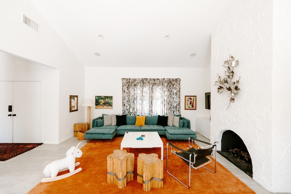 A spacious living area features an open-concept layout with a large sectional sofa accompanied by a coffee table. A bright orange area rug adds warmth, while a decorative fireplace and patterned curtains enhance the room's character. An inviting rocking horse is situated to the side.