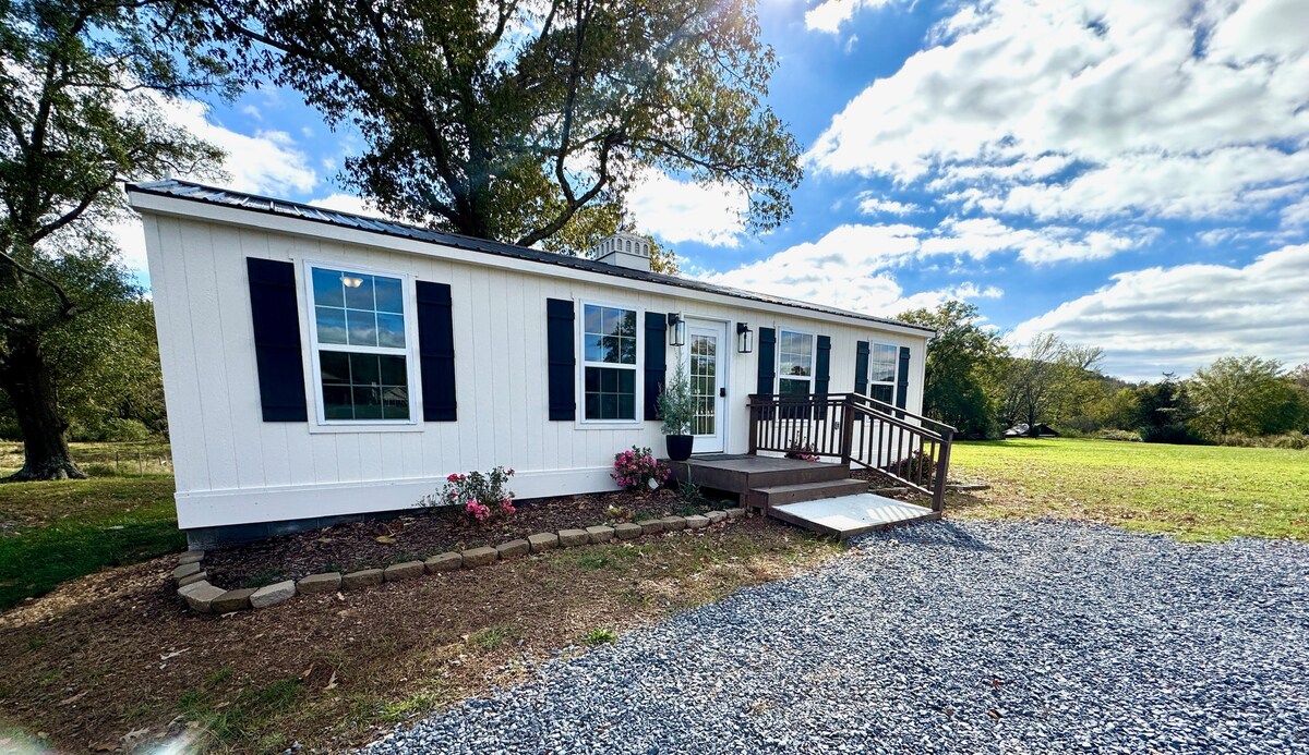 The exterior of the guesthouse is depicted, featuring a white structure with blue shutters and large windows. A wooden ramp leads to the entrance, surrounded by flower beds. A spacious lawn is visible in the background, under a partly cloudy sky.