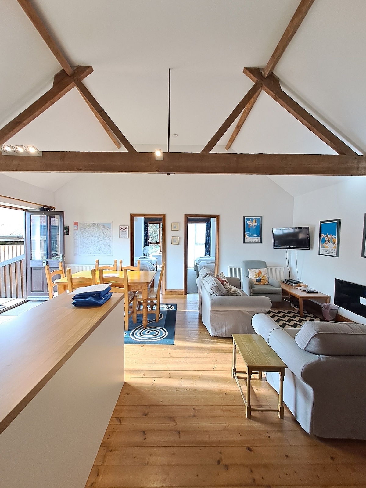 The open plan living area features a wooden ceiling with exposed beams, enhancing the spacious feel. A dining table for six is set to one side, while two sofas, a coffee table, and a TV are arranged on a cozy rug, inviting relaxation.
