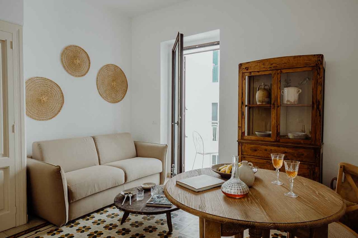 A bright living area is highlighted by a comfortable sofa and a round wooden table set for two. Decorative woven wall art is displayed, while a cabinet showcases dishware. Natural light enters through a wooden-framed window, enhancing the inviting space.