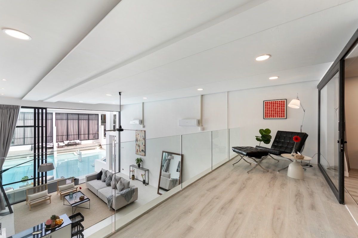 The mezzanine floor features a stylish lounge area with a sleek black chaise lounge and a small side table. A pop of color is added by a red wall art piece. Below, the open living space is visible, offering a glimpse of the pool area through large glass doors.