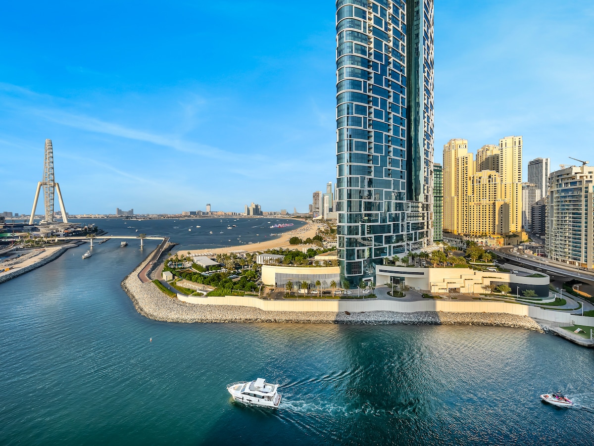 An expansive view of the coastline features a modern high-rise building alongside the waterfront. The vibrant blue water flows gently, with boats navigating while the skyline of Dubai and its sandy beaches are visible in the background, showcasing a picturesque urban landscape.