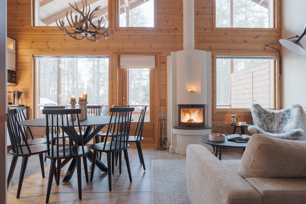 A cozy living space features a modern fireplace surrounded by natural wood accents. A round dining table accommodates six chairs, while large windows invite natural light and views of the forest. Soft furnishings include a comfortable sofa and textured throw, enhancing relaxation.