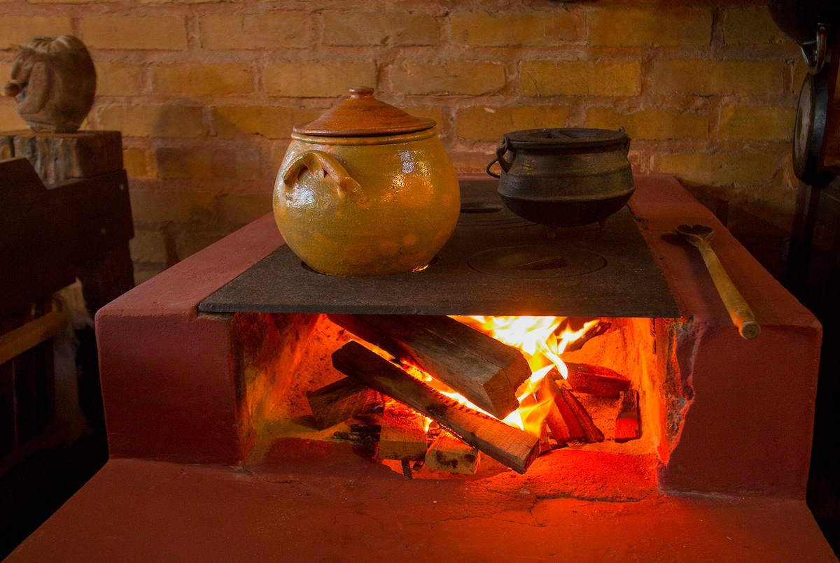 A traditional wood-fired stove is featured, with vibrant flames visible through its opening. A clay pot and a metal pot rest atop the stove, emphasizing the cooking potential. The background consists of warm, brick wall tones, creating a rustic cooking environment.