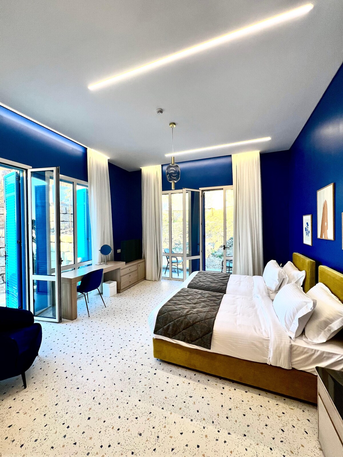 A bedroom featuring deep blue walls and large windows that open to natural light. Two spacious beds are positioned with plush headboards. A desk and chair complement the room, while contemporary artwork decorates the walls. Soft curtains frame the windows, enhancing the open atmosphere.