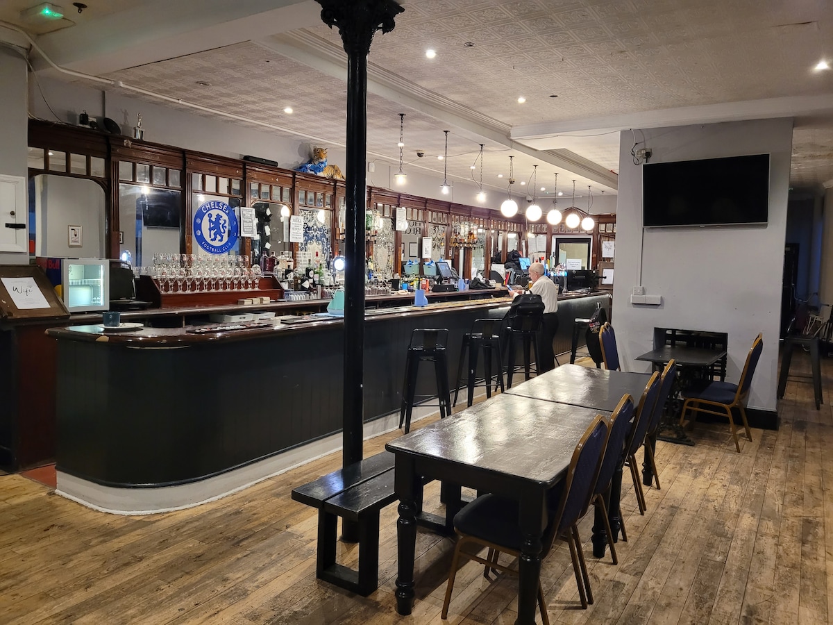 A spacious interior of a traditional pub is featured, with a long bar counter and several bar stools. Dark wooden tables are arranged throughout the area, with a wall adorned by mirrors and decorative accents. Soft lighting creates a relaxed atmosphere.