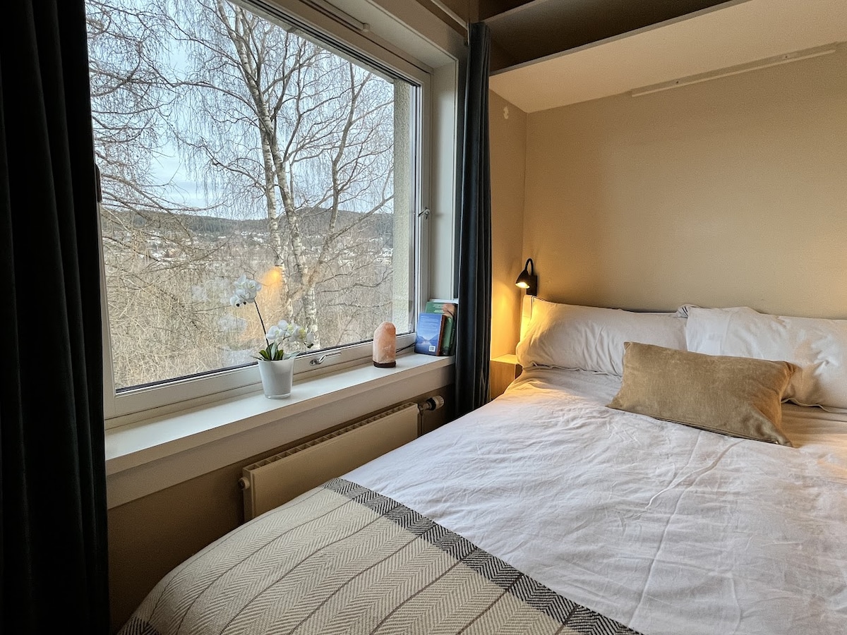 A cozy sleeping area is presented, featuring a spacious bed with neutral bedding and a decorative pillow. A large window offers a view of the surrounding landscape, while a small plant and a lamp are positioned on a nearby surface.