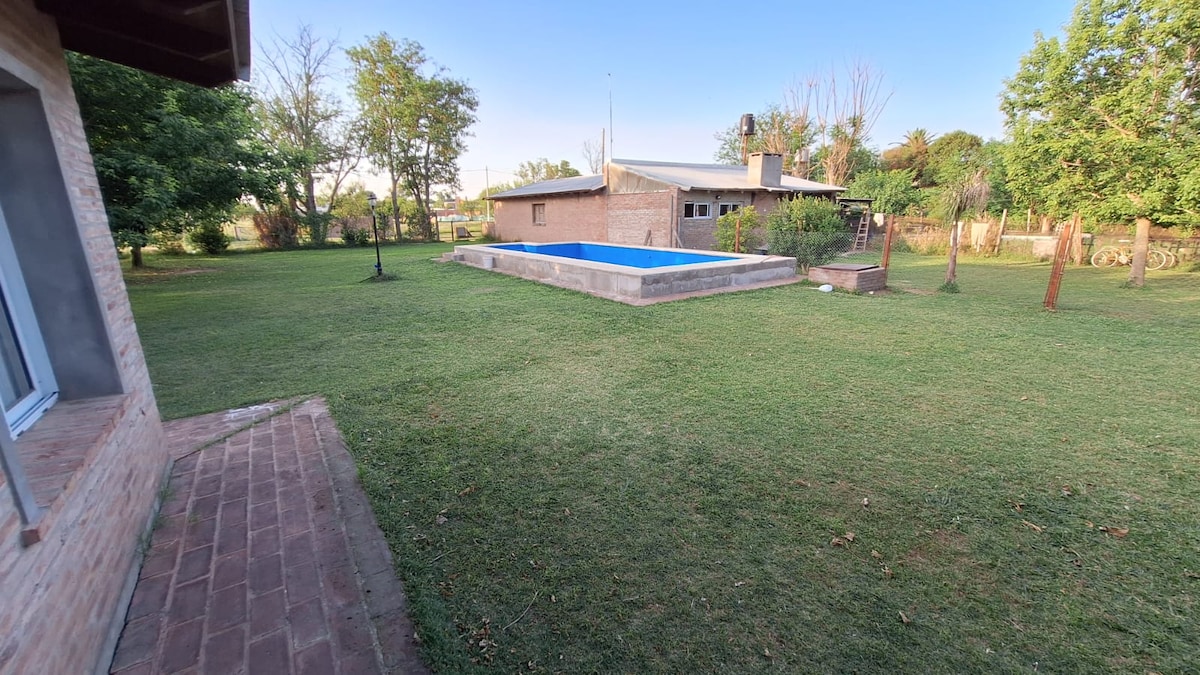 A spacious outdoor area encompasses a well-maintained lawn, featuring a rectangular blue swimming pool. Surrounding trees provide shade, and a patio area is visible, complementing the tranquil setting. The house in the background offers a simple design and contributes to the serene atmosphere.