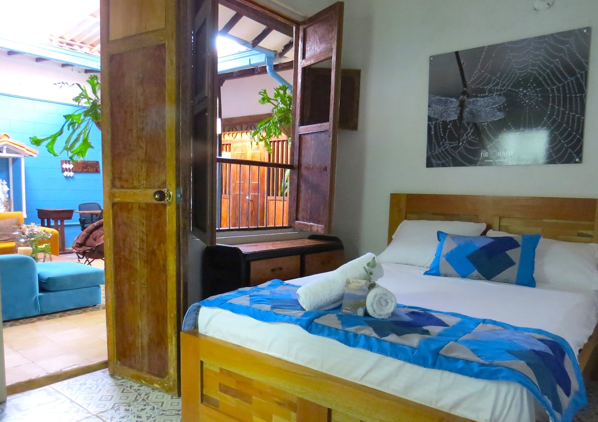 A cozy sleeping area features a wooden bed with a blue and white quilt, complemented by neatly arranged towels. Open shutters allow natural light to enter, revealing a view of a small outdoor seating area surrounded by greenery.