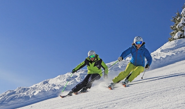 Two skiers navigate a snow-covered slope against a backdrop of clear blue skies. The individuals wear brightly colored ski outfits and helmets, showcasing the excitement of winter sports. Majestic mountains can be seen in the distance, enhancing the sense of outdoor adventure.