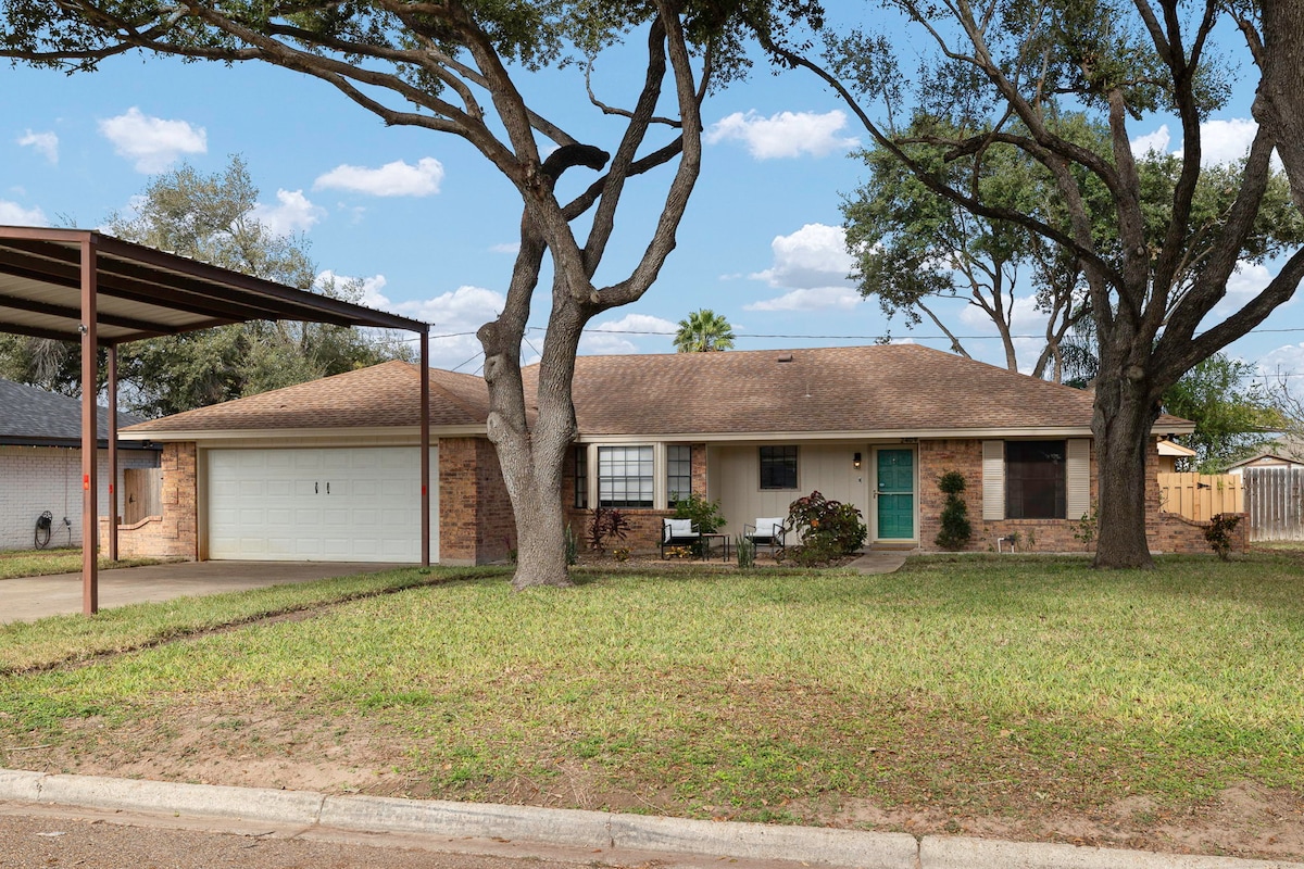 The exterior of a one-story home is showcased, featuring a covered parking area and a well-maintained lawn. Two large trees provide shade in the front yard, while a small seating area with chairs is positioned near the entrance, adding to the inviting appearance.
