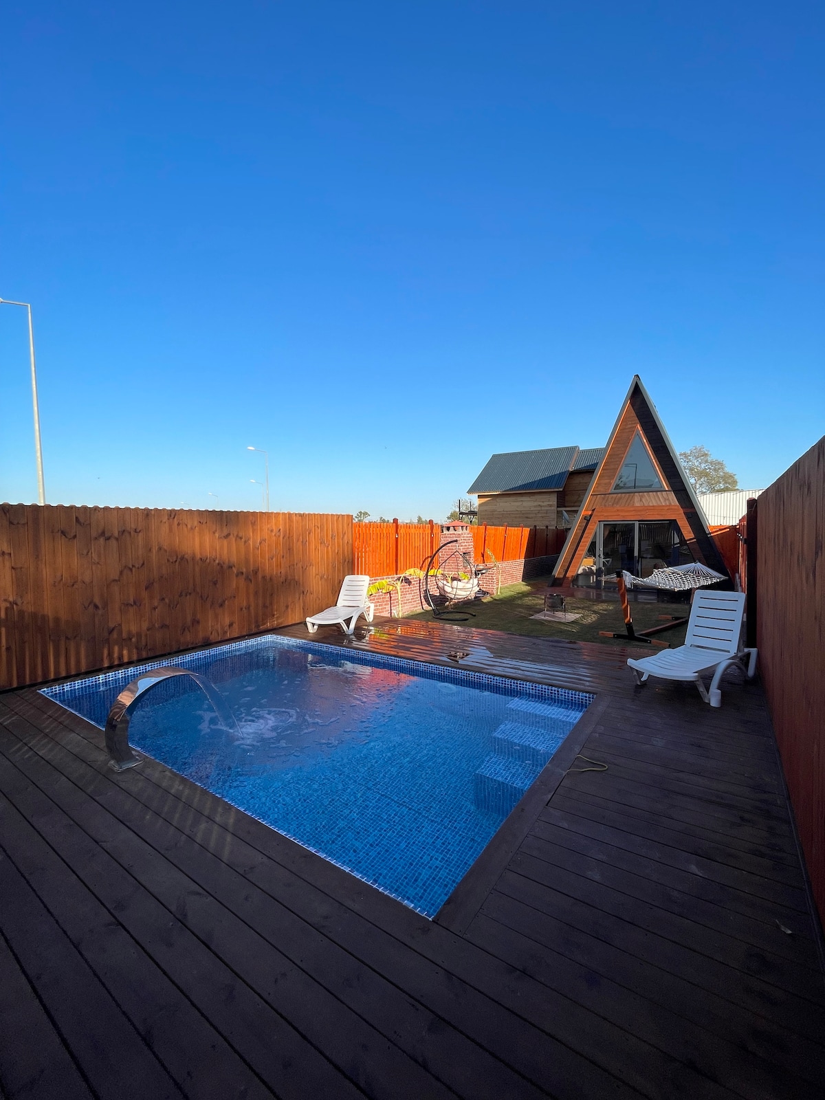 An outdoor pool with clear, blue water is surrounded by wooden decking. Comfortable white lounge chairs are positioned around the pool. A rustic wooden fence provides privacy, while a uniquely shaped cabin is visible in the background against a clear blue sky.