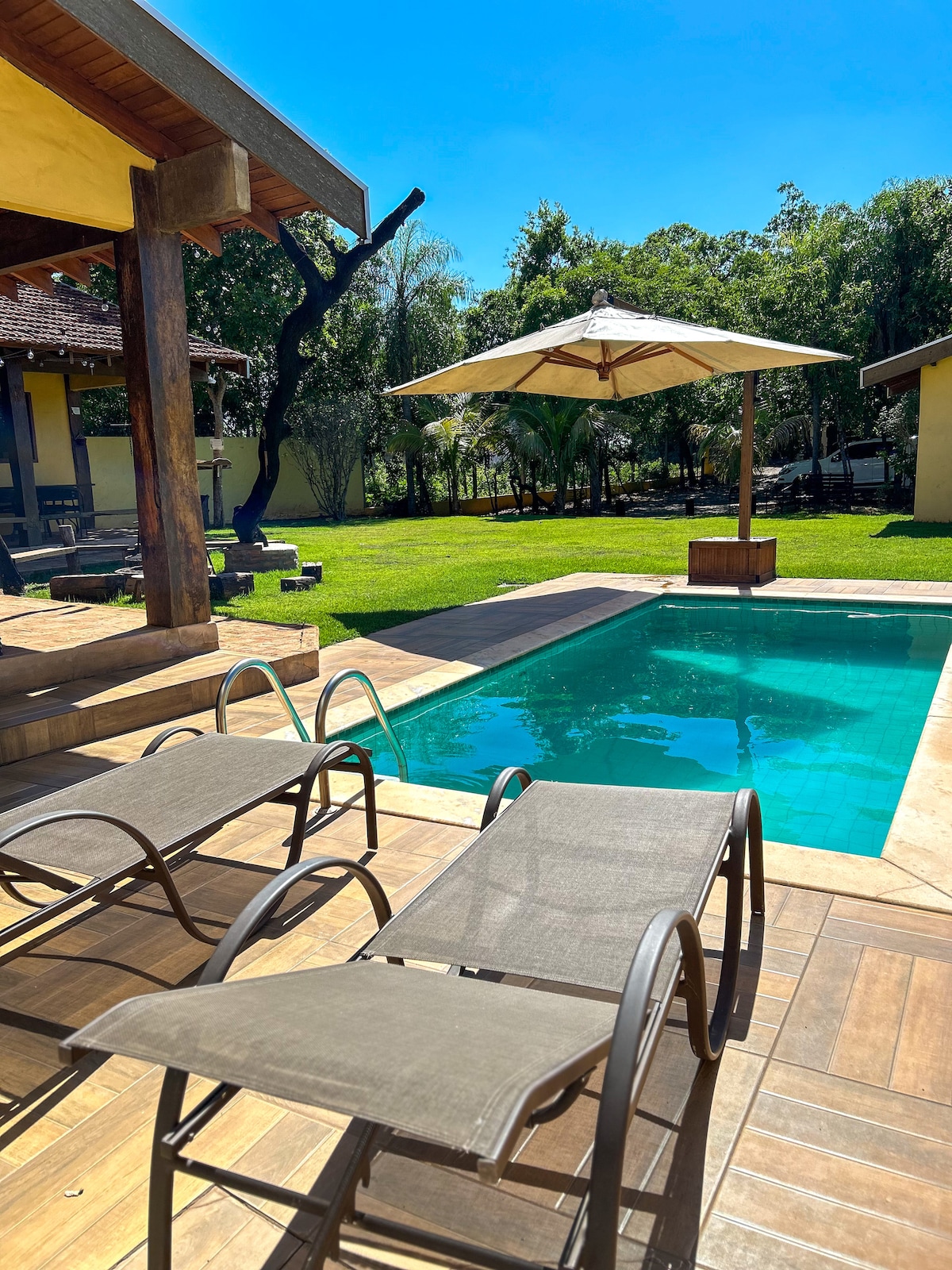 A heated swimming pool is surrounded by a well-maintained outdoor area featuring spacious loungers and an umbrella. Lush greenery frames the scene, enhancing the tranquil ambiance of the environment.