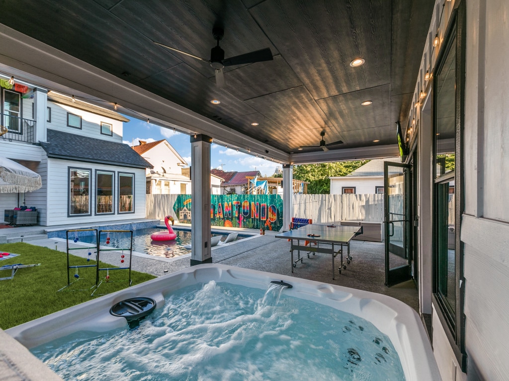 A covered patio features a heated jacuzzi, surrounded by a spacious area that includes a swimming pool and ping pong table. Colorful decorations enhance a vibrant atmosphere, while outdoor seating invites relaxation. The scene is framed by the exterior of the double home in the background.
