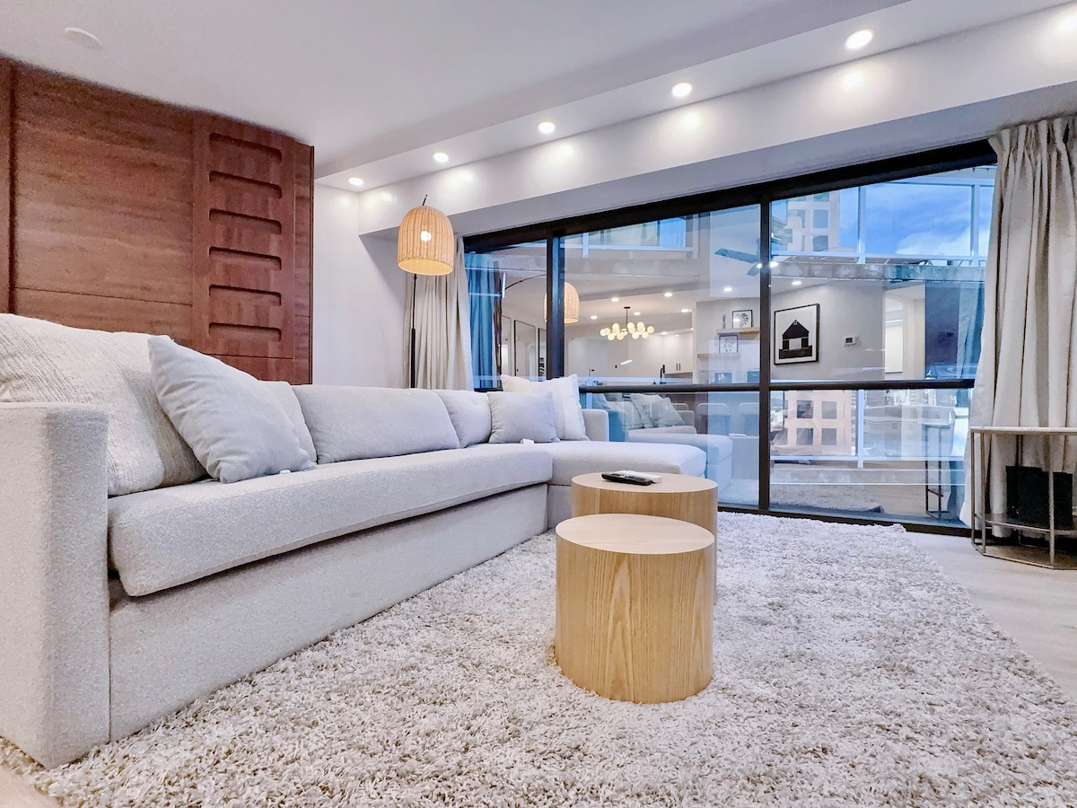 A spacious living area features a large sectional sofa, complemented by soft cushions. Two round wooden coffee tables are placed on a plush area rug. Natural light filters through the floor-to-ceiling windows, adorned with sheer curtains, revealing a view of the building's exterior.