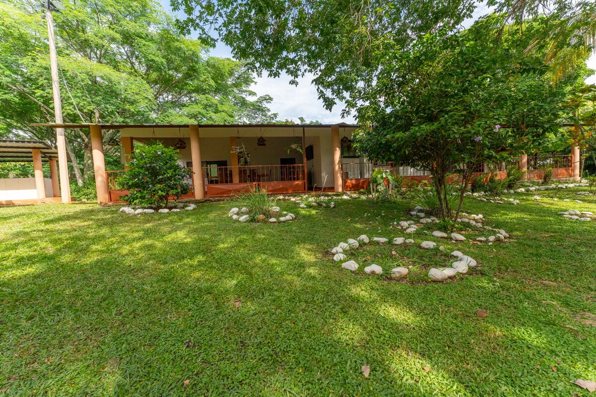 The exterior of the finca is surrounded by lush green grass and well-maintained gardens, featuring decorative stone pathways. A spacious covered porch is visible, offering shaded seating areas. Leafy trees provide a natural canopy, enhancing the relaxing outdoor environment.