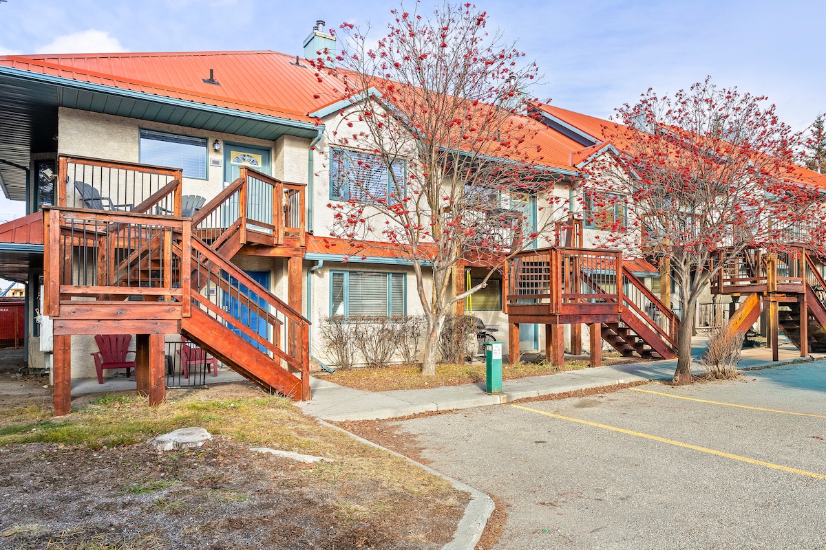 The exterior of a two-story building features a red metal roof and wooden stairs leading to the upper units. Surrounding shrubs and trees with red foliage are present, adding natural elements to the outdoor space. A small patio area with seating is visible in the foreground.