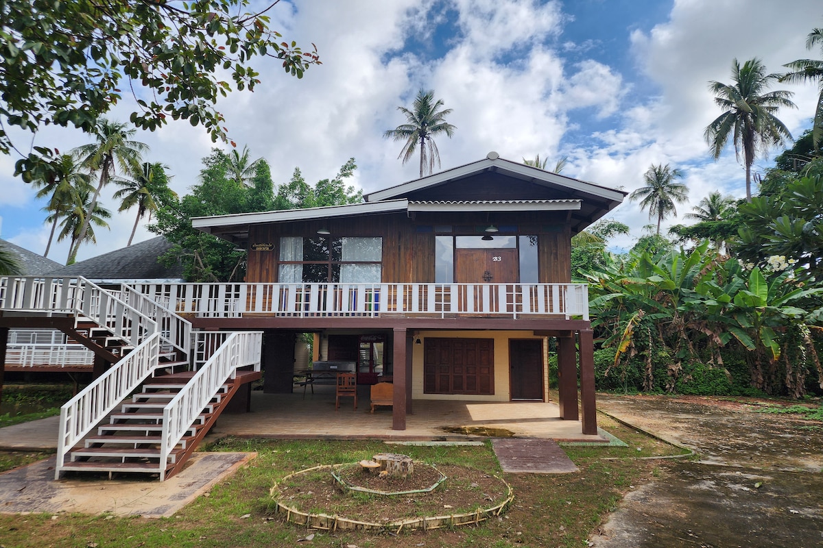 A two-story building is set against a backdrop of palm trees and blue sky. The upper level features a spacious balcony with white railings, while a set of stairs leads to the entrance. A stone fire pit is visible in the yard.