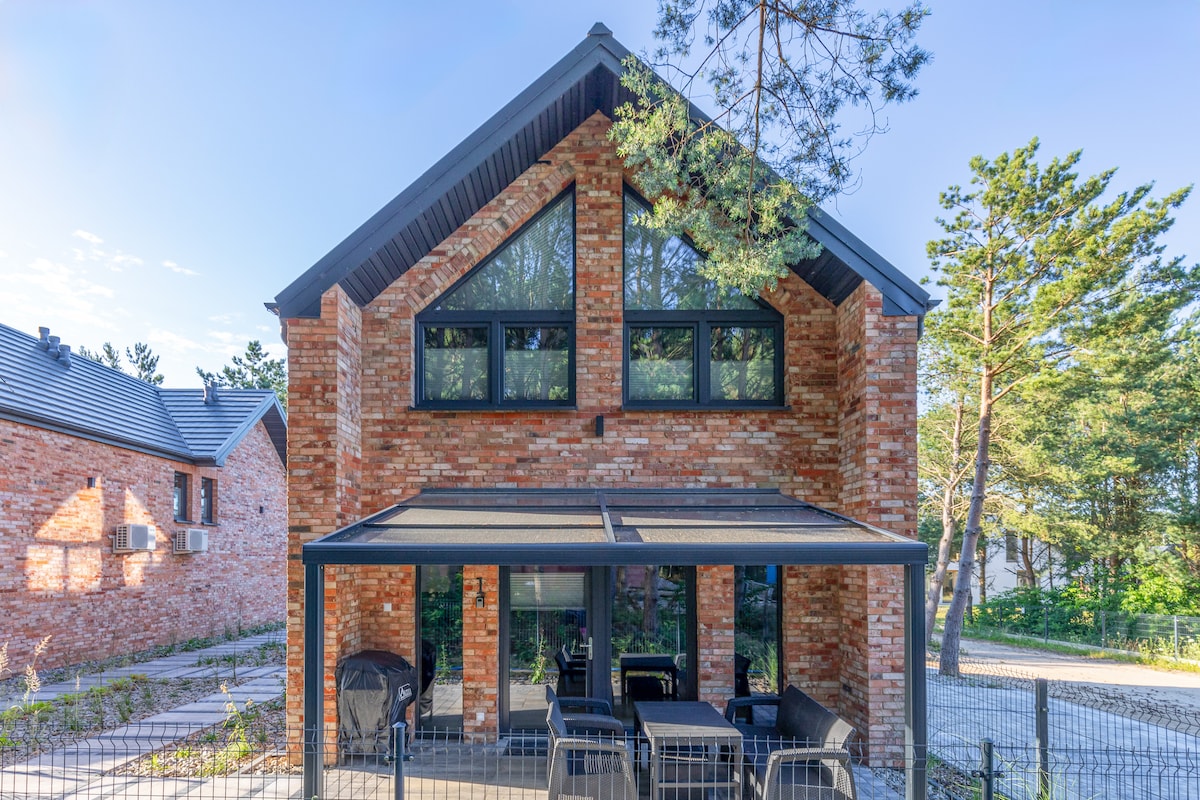 The exterior of a modern brick building showcases large windows that allow natural light to fill the space. A covered patio with outdoor seating is present, offering a relaxing area surrounded by greenery. The structure features a contemporary design with a well-maintained garden.