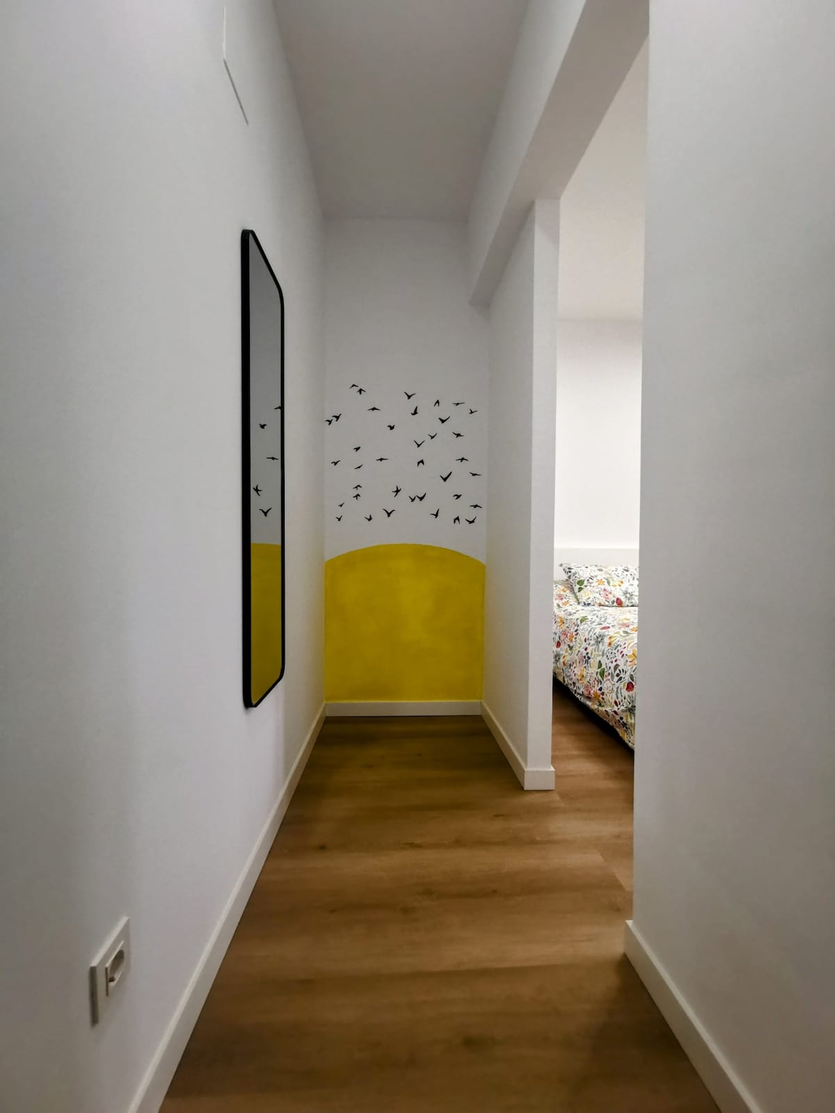 A narrow corridor is depicted, featuring a mirror on one side and a wall painted with a yellow curved shape and silhouettes of birds. At the end of the corridor, a spacious bed with colorful bedding is visible, adding a touch of brightness to the area.