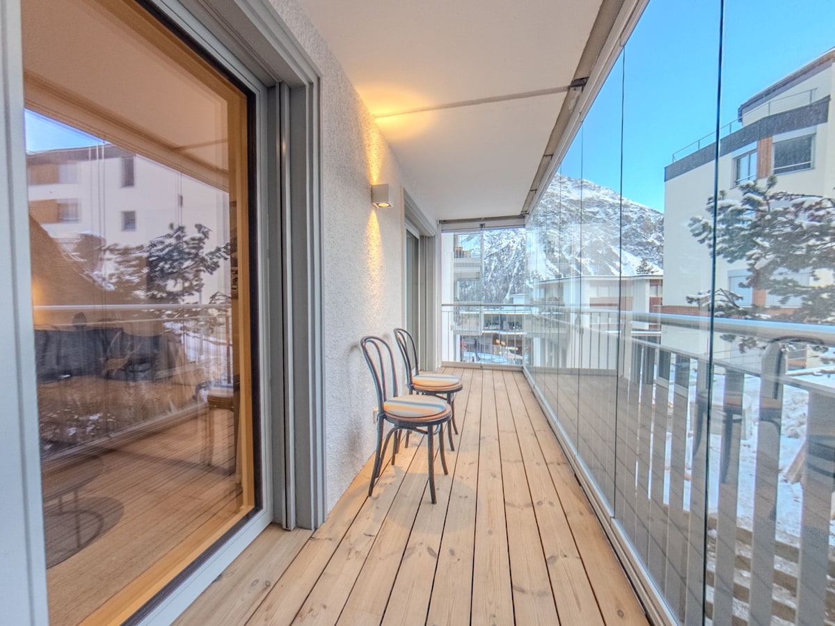 A balcony features sliding glass doors, allowing natural light to flow. Two wooden chairs with circular cushions are positioned on the wooden deck, complemented by a view of the surrounding mountains and buildings.