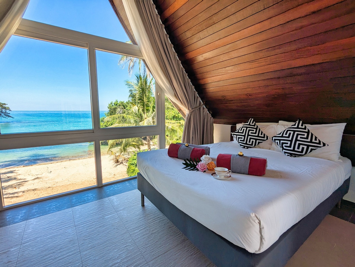 A serene bedroom features a spacious bed adorned with decorative pillows. Large glass windows provide a stunning view of the beach and sea, while soft curtains frame the scene, allowing natural light to enhance the room's ambiance. An inviting atmosphere is created by the coastal setting.