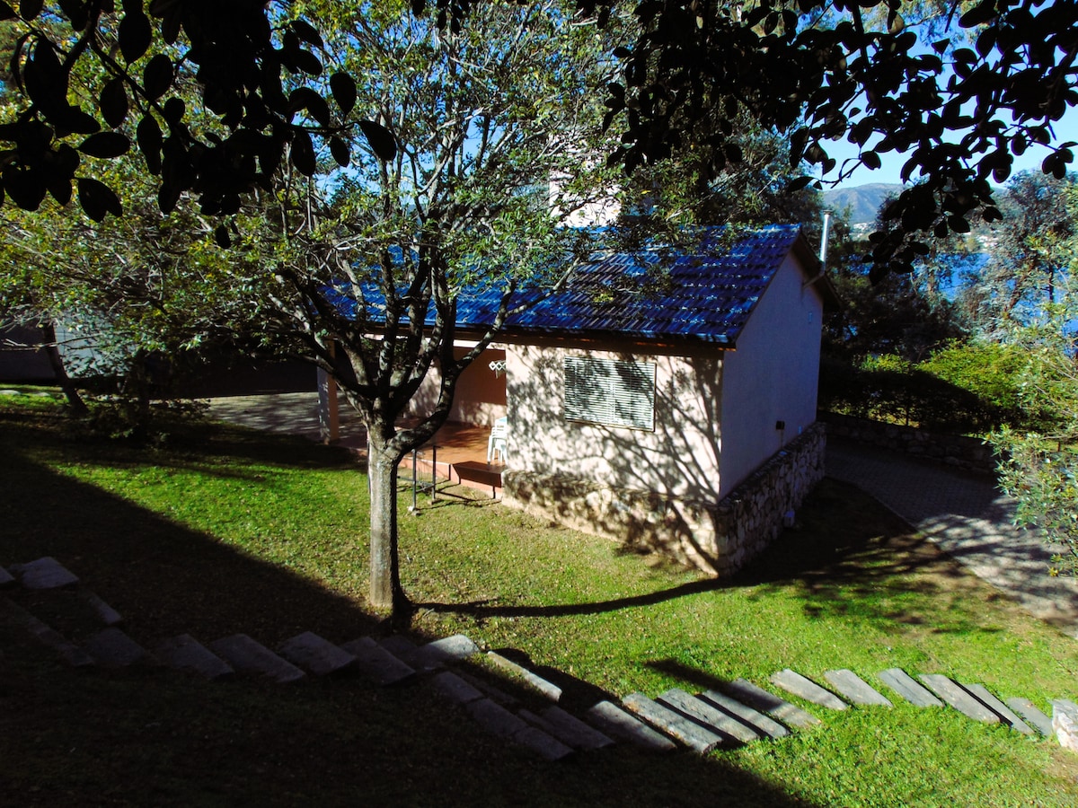 A rustic cabin is nestled in a lush green landscape, featuring a stone pathway leading to the entrance. The structure is framed by trees and natural foliage, with a blue-tiled roof and light-colored walls. The serene atmosphere is complemented by views of the nearby lake.